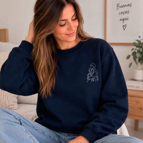 Mujer joven con sudadera azul marino que tiene un bordado lineal de una pareja abrazada, sentada en una habitación con decoración minimalista.